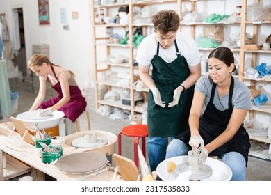 Young woman teacher in apron helps students teenage girl and teenage boy sculpt ceramic product from raw clay on potter's wheel - Powered by Shutterstock