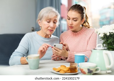 Young woman teaching her aged mother to use smartphone while sitting at table in living room.. - Powered by Shutterstock