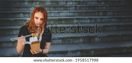 young woman or teenage girl eating asian fast food from takeaway box sitting on the steps on city street. Thai noodles in paper box takeaway street food