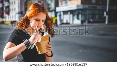 young woman or teenage girl eating asian fast food from takeaway box on city street. Thai noodles in paper box takeaway street food