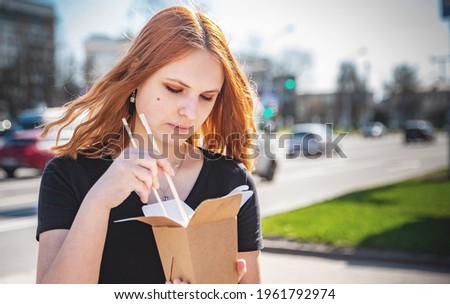 young woman or teenage girl eating asian fast food from takeaway box on city street. Thai noodles in paper box takeaway street food