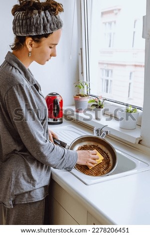 young woman, teenager, daughter, in gray pajamas, with bandage on brown hair, washes dishes in sink in small modern kitchen. teenager washes burnt frying pan in kitchen with sponge in hand.