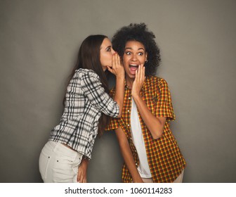 Young woman telling her girlfriend some secret. Two women gossiping. Excited emotional girl whispering to her friend ear, gray studio background