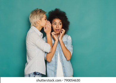 Young woman telling her girlfriend some secret. Two women gossiping. Excited emotional girl whispering to her friend ear, turquoise studio background