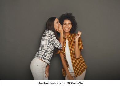 Young woman telling her girlfriend some secret. Two women gossiping. Excited emotional girl whispering to her friend ear, gray studio background