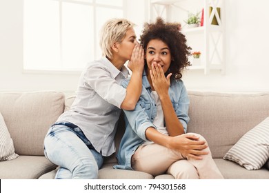 Young woman telling her girlfriend some surprised secret. Two women gossiping. Excited emotional girl whispering to her friend ear while sitting in living-room