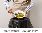 Young woman throwing organic food into trash bin on grey background, closeup. Waste recycling concept