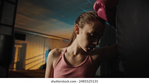 Young woman throwing a powerful punch at a heavy bag in a dimly lit gym with an illuminated boxing ring, showcasing intense training and fitness dedication - Powered by Shutterstock