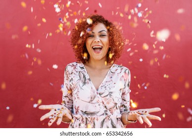 Young Woman Throwing Star Shaped Colorful Confetti In Air. Close Up Of Woman In Happy Mood With Confetti All Around Against A Red Background.