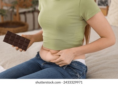 Young woman in tight jeans with chocolate sitting on bed at home, closeup. Weight gain concept - Powered by Shutterstock