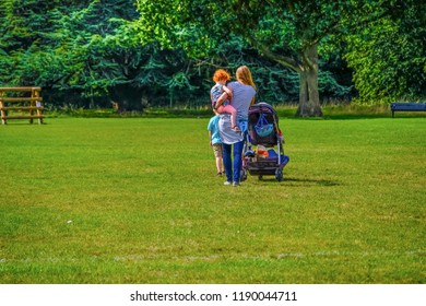 Young Woman With A Toddler And Older Boy Walking In The Park And Pushing Stroller During The Day