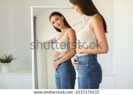 Young Woman Touching Flat Belly Expecting Baby And Enjoying First Months Of Pregnancy Posing Looking At Her Reflection In Mirror Wearing Skinny Jeans Standing At Home. Selective Focus