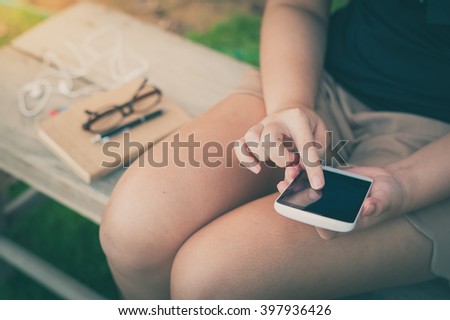 Young woman touching on smartphone screen while sitting on wood bench in park in morning time on weekend with vintage filter effect