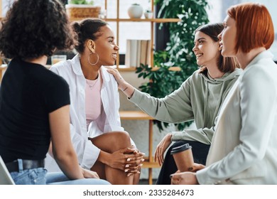 young woman touching shoulder of pleased african american girlfriend while sitting next to redhead psychologist with paper cup during therapy in consulting room, mental wellness concept - Powered by Shutterstock