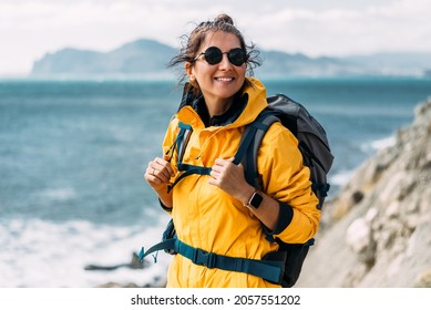 A young woman in tourist gear stands against the background of the sea and looks into the camera. A traveling woman with a backpack smiles at the camera. Portrait of a smiling tourist. Smiling tourist