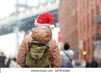 A Young Woman Tourist In A Santa Claus Hat Walks During A Snowfall In New York On Christmas Eve. Manhattan Bridge And Skyscrapers Of NY On Snowy Day. Winter Holidays In NYC. Snow Is Rare In New York.