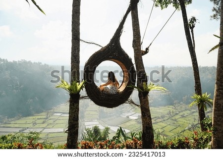 A young woman tourist sitting in a bird nest, immersed in the breathtaking green landscape of Bali on a sunny day. Tourist doing jungle swing. Rice fields and forest in background. Indonesia.