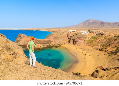 Young Woman Tourist Standing On Cliff Looking At Papagayo Beach, Lanzarote, Canary Islands, Spain