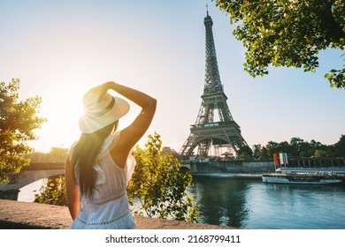 Young woman tourist in sun hat and white dress standing in front of Eiffel Tower in Paris at sunset. Travel in France, tourism concept