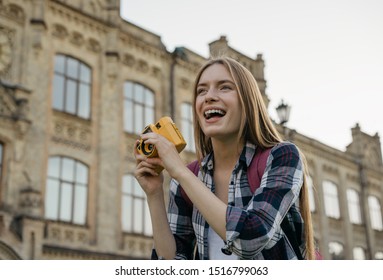 Young Woman Tourist Taking Photo On Retro Camera. Portrait Of Professional Photographer Holding Yellow Camera. Happy Hipster Traveler With Backpack Walking On Street, Laughing   