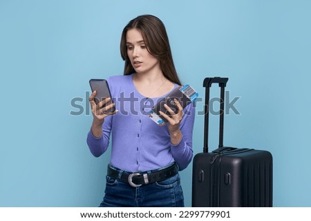 Young woman tourist using smartphone, checking mobile app, online registration of flight, holding boarding pass, posing with suitcase over blue background. Travel Tourism Journey People Technology