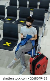 Young Woman Tourist Wearing Face Mask Holding Passport And Ticket, Sitting In Airport Or Train Station On Empty Marked Chairs Under New Normal And Social Covid Distance Regulations