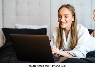 Young woman in towel and bathrobe sitting on bed at home and working on her laptop. Beautiful young woman wearing a white bathrobe, with a towel on her head in bed at home - Shutterstock ID 1920061985