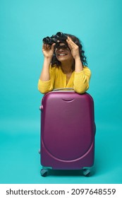 Young woman traveler adventurer explorer tourist in bright yellow clothes leaning on a suitcase and smiles toothy smile looking through binoculars, isolated on blue background with copy space