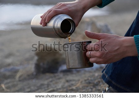 young woman traveler hands pouring tea from a thermos in an iron mug, near the fire on the coast, close up. drinking tea during hike. camping concept