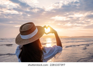 Young woman traveler making heart shape with hands and looking beautiful sunset on the beach, Travel lifestyle concept