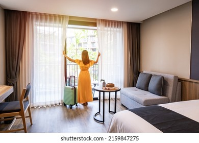 Young woman traveler opening the curtains and looking at the view from the window of a hotel room while on summer vacation, Travel lifestyle concept