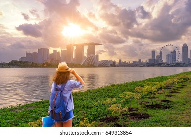 Young woman traveler with sky blue backpack and hat holding the map with singapore city downtown background. Traveling in singapore at sunset times