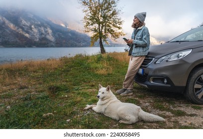 Young woman traveler standing next to the car while traveling with her white swiss shepherd dog on the shore of a mountain lake in foggy autumn weather - Powered by Shutterstock