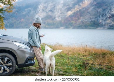 Young woman traveler standing next to the car while traveling with her white swiss shepherd dog on the shore of a mountain lake in foggy autumn weather - Shutterstock ID 2240300951