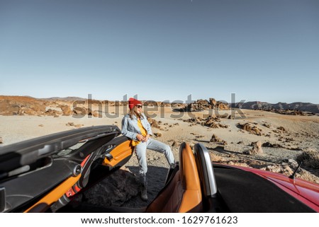 Young woman traveling by convertible sports car, standing with map on the roadside of the desert valley. Wide landscape view