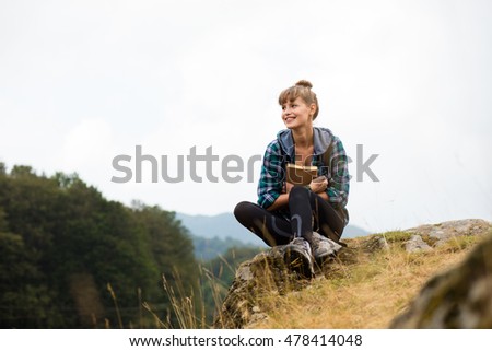 young woman traveling with her backpack in the mountains, sitting on a rock and holding a book or a journal in hands thinking about something
