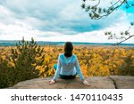 Young woman traveller sitting on a steep rock, admiring panoramic view of autumn forest in Algonquin Provincial Park, Ontario, Canada. Solo travelling, rear view