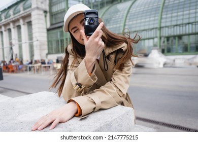 Young Woman In Trench Coat And Baseball Cap Taking Photo On Vintage Camera