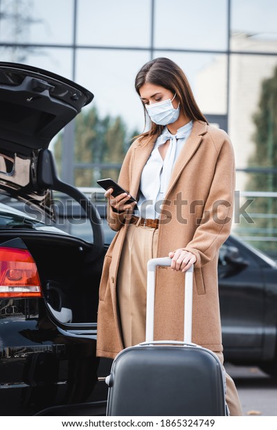 young woman in\
trench coat and medical mask messaging on smartphone while standing\
with suitcase near car\
trunk