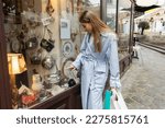 young woman in trench coat standing with shopping bags and pointing at showcase of antique shop in Vienna