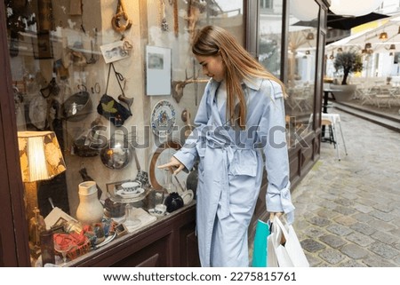 young woman in trench coat standing with shopping bags and pointing at showcase of antique shop in Vienna