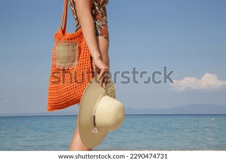 Young woman in tropical printed swimwear and straw hat cary orange crochet tote bag empty sandy beach on, summer fashion style, relaxed vacation	