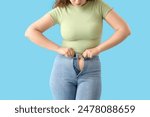 Young woman trying to button tight jeans on blue background, closeup. Weight gain concept