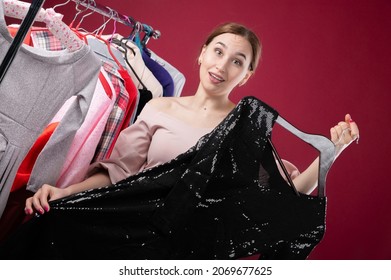 Young Woman Trying To Choose A Dress To Wear, Holding Black Clothes On A Hanger With A Hesitant Look, Trying To Make A Decision