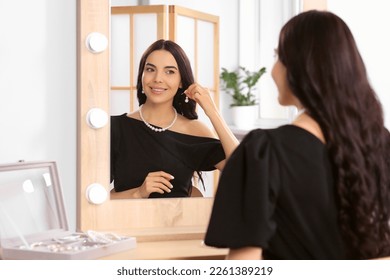 Young woman trying on elegant pearl earring near mirror indoors