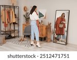 Young woman trying on stylish clothes with bag near mirror at home