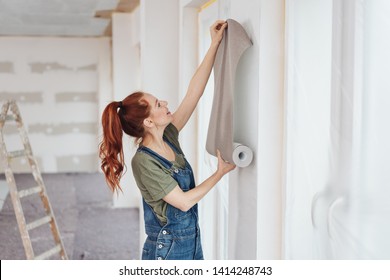 Young Woman Trying Out New Wallpaper At Home Holding The Roll Up Against A Newly Painted White Wall For Visual Effect