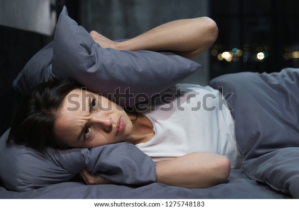 Young woman trying to sleep but\
disturbed by noisy neighbors and covering ears with\
pillows
