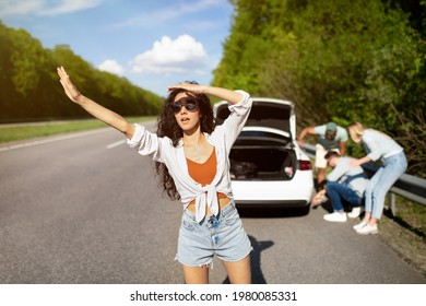 Young woman trying to wave down car, asking for help, her friends trying to fix their broken vehicle, having flat tire on roadside. Millennial lady hitchhiking on highway, needing assistance - Powered by Shutterstock