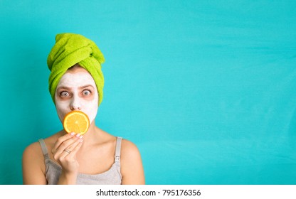 Young Woman With A Turban Of Green Towels With A White Mask On Her Face Holds Near The Mouth Of An Orange And Is Very Surprised On A Blue Background. Popart Woman. Pop Art